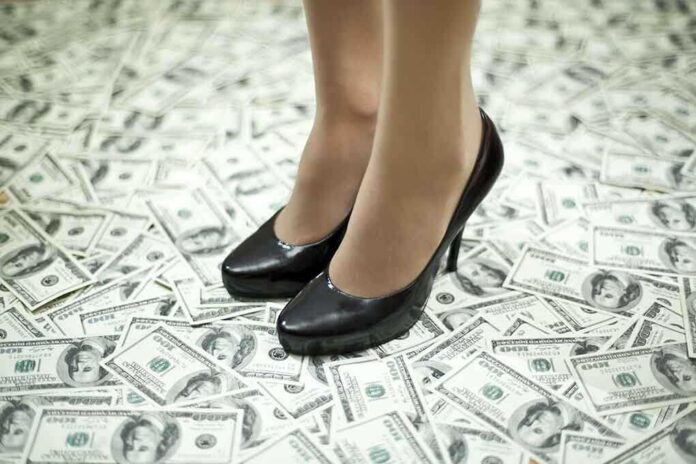 A pair of black high heels standing on a floor covered with dollar bills