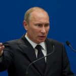 A man in a suit gesturing while speaking at a podium with microphones against a blue background