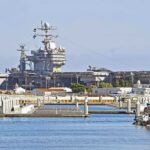A large naval aircraft carrier docked in a harbor with smaller boats in the foreground