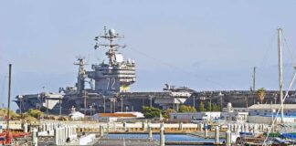 A large naval aircraft carrier docked in a harbor with smaller boats in the foreground