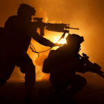 Two soldiers with weapons silhouetted against fiery background.