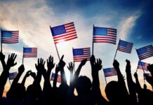 A National Milestone Becomes a Political Battleground Silhouetted crowd holding American flags against a sunset sky