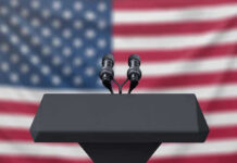 Podium with microphones, American flag background.