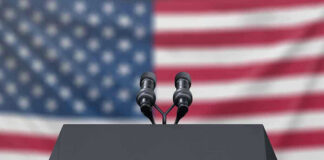Podium with microphones, American flag background.