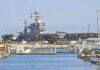 Defense Startup Wins Contract for Long-Range A large naval aircraft carrier docked in a harbor with smaller boats in the foreground