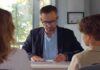Man in glasses reviewing papers at a desk with two people