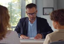 Man in glasses reviewing papers at a desk with two people