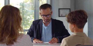 Man in glasses reviewing papers at a desk with two people