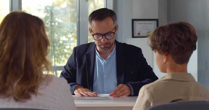 shutterstock_2041483871 Man in glasses reviewing papers at a desk with two people
