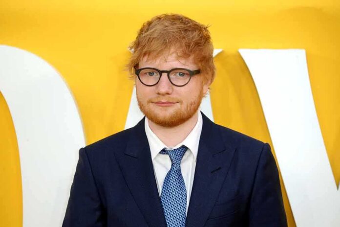 shutterstock_1946174674 Ed Sheeran in a navy suit against a yellow backdrop