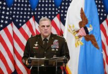 Military officer speaks at podium with U.S. flags behind