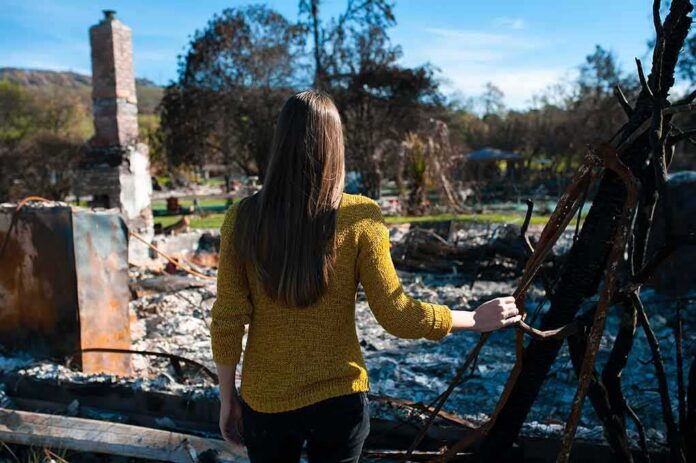 shutterstock_1141486334 Woman in yellow sweater surveys burned ruins outdoors