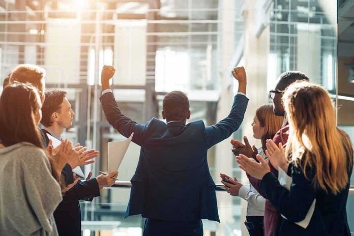 Businessman raising fists as colleagues applaud in office
